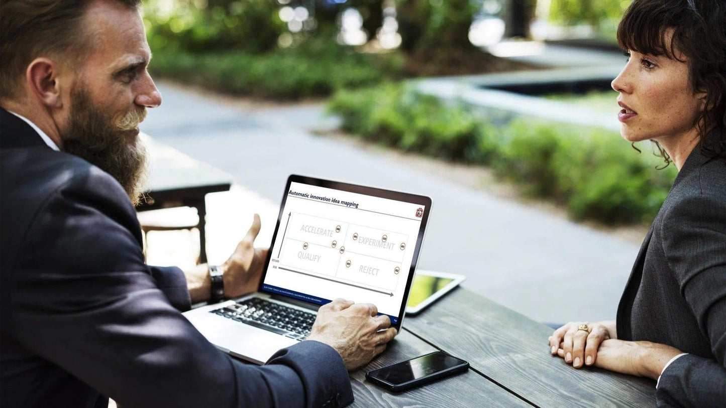 A man and a woman are sitting at an outdoor wooden table, deep in conversation. The man gestures towards a laptop showcasing the Innovation Prioritization Tool & Dashboard, which focuses on accessibility, speed, quality, and reset options. Next to a smartphone on the table is an Expert Toolkit. In the background are blurred greenery and a sidewalk.