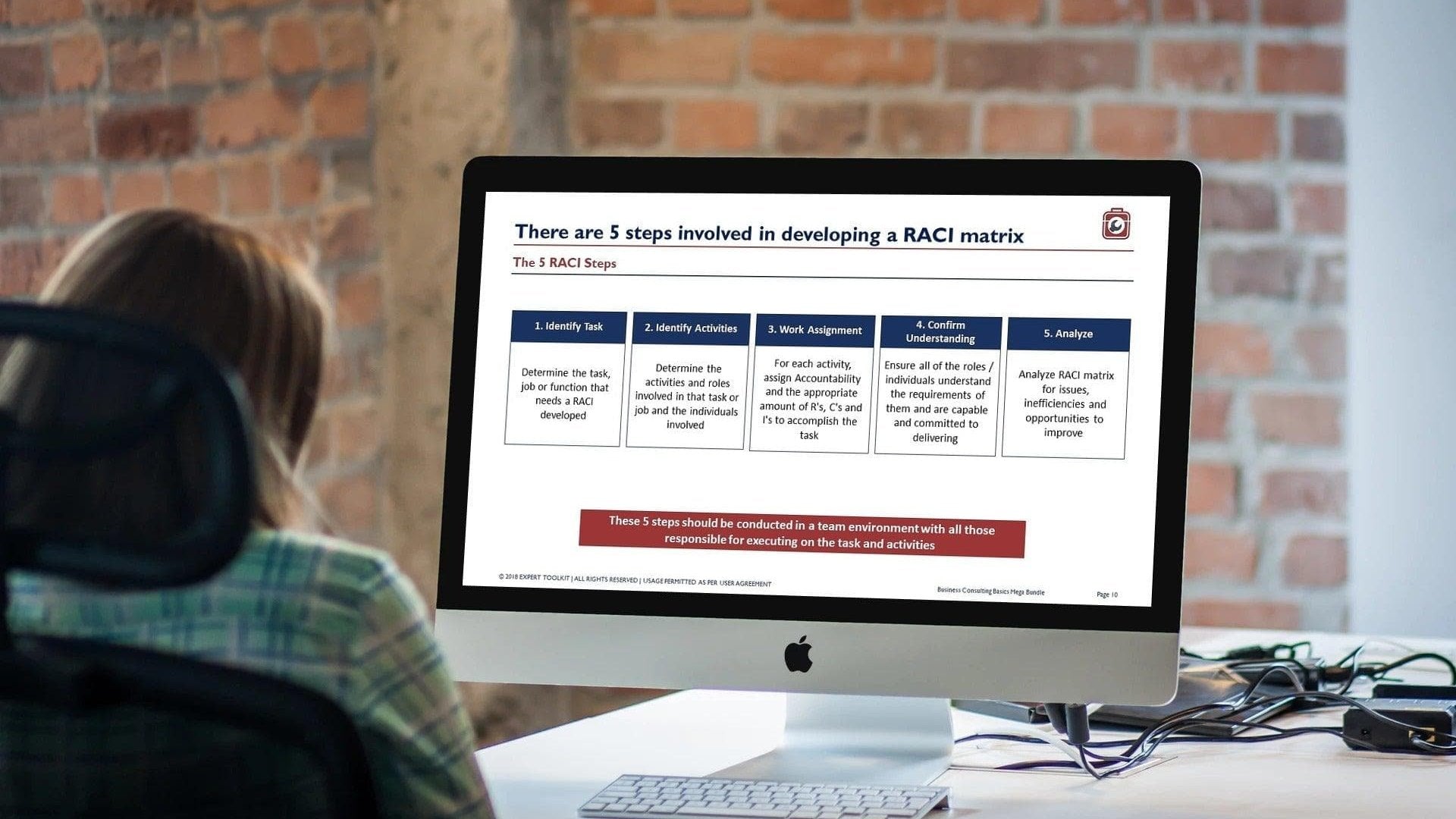 A person is seated at a desk, examining a computer screen that showcases the "Management Consulting Basics" presentation. This particular slide emphasizes creating a RACI matrix and details the five essential steps with comprehensive business analysis skills. An exposed brick wall is featured as part of the office's backdrop in this setting.