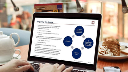 A person is utilizing "The Master Change Management Practitioner" laptop placed on a table, displaying a document titled "Preparing for Change." The document includes a flowchart that covers aspects such as Change Readiness Assessment, Stakeholder Mapping & Engagement, and Action Plan. Accompanied by coffee and cake, they immerse themselves in change management strategies for clarity using tools like the RACI Matrix.