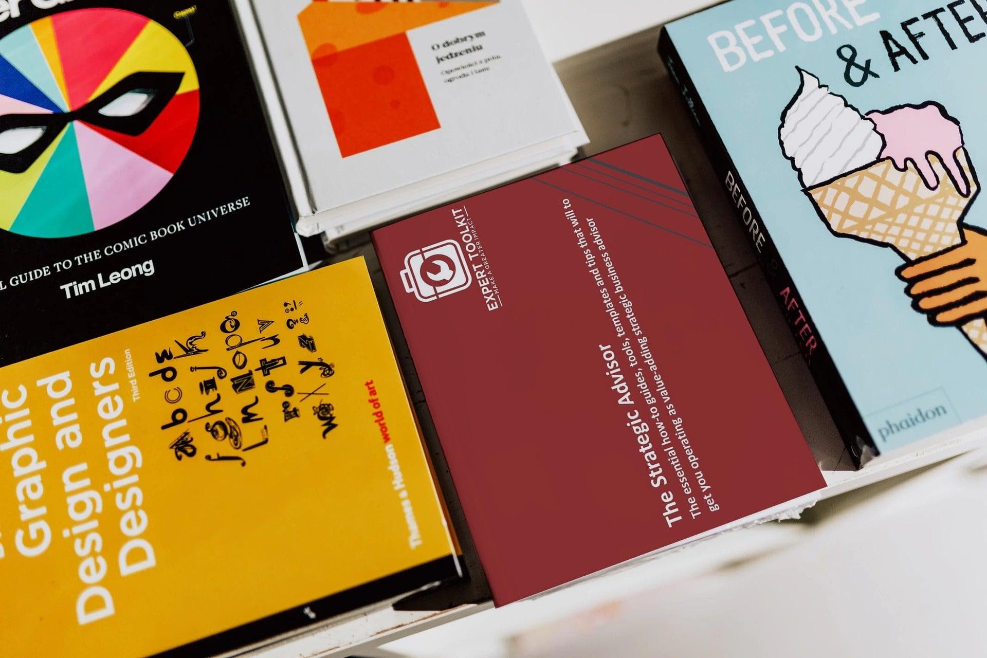 On a white surface lies a collection of books, including "Graphic Design and Designers" adorned with a colorful circle. Among them is "The Strategic Advisor" from Purchase Only | No Online Access, offering valuable insights for business consultants. Meanwhile, "Before & After" showcases an ice cream cone graphic. Other books in the collection feature abstract patterns and text designs.